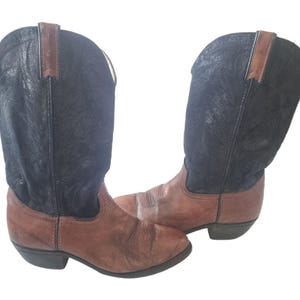 Könnte beinhalten: Ein Paar Cowboy-Stiefel mit klassischem Design. Die Stiefel haben braune Lederfußbetten und schwarze Schäfte mit einem strukturierten Muster. Die Stiefel haben eine traditionelle spitze Zehenpartie und einen gestapelten Absatz.