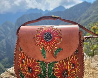 May include: A brown leather crossbody bag with a flap closure. The bag features hand-painted sunflowers in red, yellow, and green. The bag has a brown strap and a gold-colored buckle. The bag is sitting on a rock.