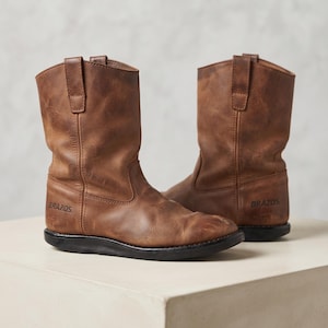 May include: A pair of mid-calf brown leather boots with a distressed finish, featuring pull tabs at the top and the word BRAZOS embossed on the outer side. The boots have a rounded toe, visible stitching along the shaft, and a solid black sole.