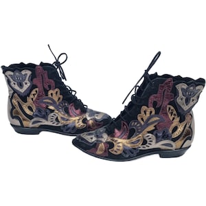 Puede incluir: Botines negros con intrincados bordados florales en dorado, burdeos y plateado. Los botines, con cordones, presentan un tacón bajo y un diseño vintage.