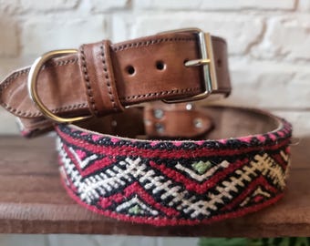 May include: Two dog collars. The top collar is brown leather with a silver buckle and gold-colored D-ring. The bottom collar is a woven design with red, white, and green geometric patterns. Both collars are on a wooden surface.