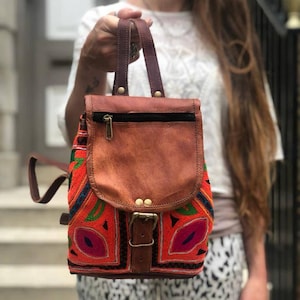 May include: A small, colorful backpack with a brown leather flap and straps. The front of the bag features intricate embroidery in red, orange, green, and blue. The bag has a buckle closure and a zipper pocket.