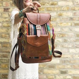 May include: A brown leather backpack with a colourful woven fabric panel. The backpack has a flap closure, a front pocket, and adjustable straps. The bag is held up by a person.