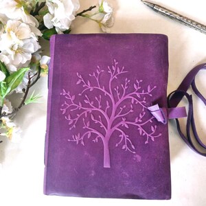 May include: A purple journal with an embossed tree design on the cover. It features a matching ribbon closure and a long strap. A silver pen and white flowers are also in the image.
