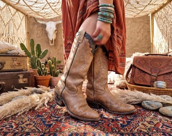 Könnte beinhalten: Abgenutzte, hellbraune Leder-Cowboystiefel mit dekorativen Nähten. Dazu ein rostfarbenes Kleid und türkisfarbene Armbänder. Die Szene spielt sich auf einem gemusterten Teppich mit einer Ledertasche und einem dekorativen Schädel im Hintergrund ab.
