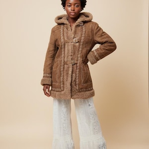 May include: A brown suede coat with a hood and decorative stitching. The coat features a shearling lining and buckle closures. The model is wearing white lace pants and black boots. The coat is a vintage style.