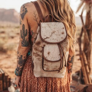 May include: A backpack crafted from white and brown cowhide with a textured hair-on-hide finish. The bag features brown leather trim, a top flap with a magnetic closure, a front pocket with a drawstring, and adjustable brown leather shoulder straps.