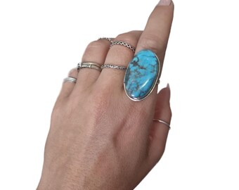 May include: A hand adorned with multiple rings, including a prominent oval turquoise ring with a silver surround. Additional rings feature intricate designs. The rings are presented against a white backdrop.