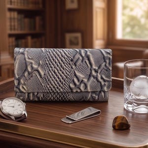 May include: A snakeskin-effect wallet, a wristwatch with a brown leather strap, a silver money clip, a glass with a spherical ice cube, and a small stone on a wooden tray.