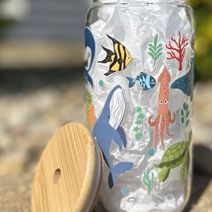 May include: A clear glass jar with a bamboo lid, decorated with colorful sea creature decals. The jar features a whale, fish, octopus, and turtle. The lid is round and has a hole in the center. The jar is filled with white tissue paper.