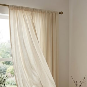 Peut inclure: Rideaux en lin beige suspendus &agrave; une tringle dor&eacute;e. Les rideaux sont partiellement ouverts, r&eacute;v&eacute;lant une fen&ecirc;tre et la verdure ext&eacute;rieure. Le tissu a une texture douce et fluide, cr&eacute;ant une sensation de l&eacute;g&egrave;ret&eacute; et d'a&eacute;ration. Les rideaux ajoutent une touche d'&eacute;l&eacute;gance.
