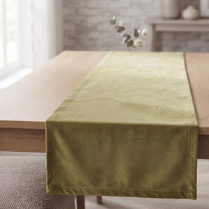 Puede incluir: Un camino de mesa rectangular hecho de tela de terciopelo color verde oliva. El camino presenta una superficie suave y lisa con un borde cosido a lo largo de los lados, extendido a lo largo del centro de una mesa de madera clara.
