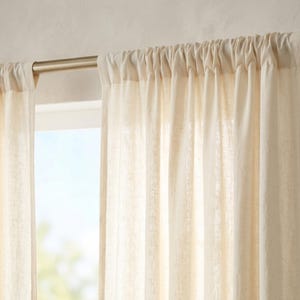 Peut inclure: Rideaux en lin cr&egrave;me suspendus &agrave; une tringle en nickel bross&eacute;. Les rideaux sont fronc&eacute;s en haut, cr&eacute;ant des plis souples. La lumi&egrave;re naturelle filtre &agrave; travers la fen&ecirc;tre derri&egrave;re les rideaux, illuminant le tissu.