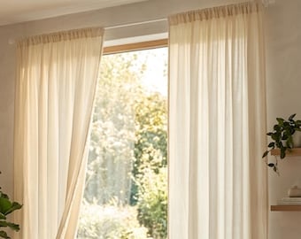 Peut inclure: Des rideaux transparents de couleur crème sont suspendus devant une fenêtre, partiellement ouverts pour révéler une scène extérieure ensoleillée et lumineuse. Les rideaux sont en tissu léger et sont suspendus à une simple tringle. La pièce est décorée de plantes.