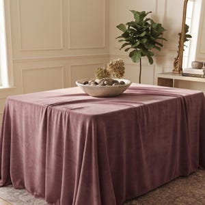 May include: A rectangular table covered with a solid dusty rose velvet tablecloth that drapes to the floor. Centered on the table is a large, shallow, off-white bowl filled with smooth grey river stones and dried beige hydrangea flower heads.