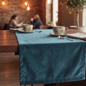 Peut inclure: Un chemin de table rectangulaire en velours bleu sarcelle avec des bords cousus, posé sur une table en bois. Deux tasses en céramique à motifs mouchetés reposent sur des soucoupes assorties, avec une cuillère en argent et une serviette beige.