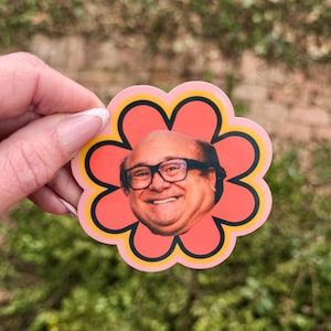May include: A sticker featuring a smiling face of a man with glasses, set within a flower shape. The flower has a coral-colored center, a yellow outline, and a black border. The background is blurred green.
