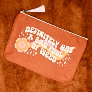 May include: Orange canvas zippered pouch with the text "DEFINITELY NOT A BAG FULL OF WEED" in white, with a retro flower and rainbow design. The pouch has a white zipper and a white tassel.