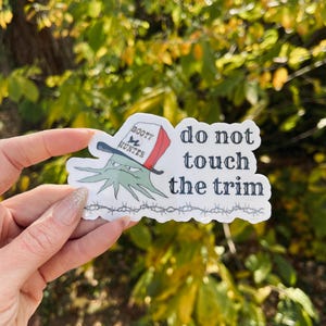 May include: A sticker with the text "do not touch the trim" and a cartoon octopus wearing a hat that says "Booty Hunter". The sticker has a barbed wire design at the bottom and is held against a background of green leaves.