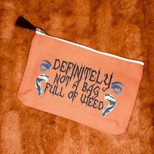 May include: Orange canvas zippered pouch with the text "DEFINITELY NOT A BAG FULL OF WEED" in black gothic lettering. The pouch has a white zipper and a black tassel. The design includes blue eyes and open mouths.
