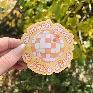 May include: A round sticker with a disco ball design in orange and white, surrounded by the text "so much panic & a little disco" in orange script. The sticker has a light orange border and star accents.