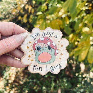 May include: A sticker featuring a cartoon mushroom character with a smiling face and the text "I'm just a fun lil guy." The mushroom has a red cap with white spots and is set against a light beige background with gold star accents.