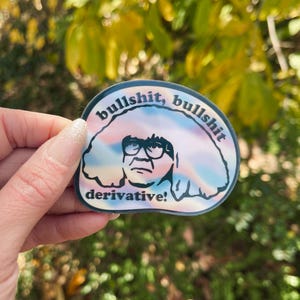 May include: A sticker featuring a cartoon image of a man with glasses, with the words "bullshit, bullshit" and "derivative!" in a colourful, swirling design. The sticker is held by a hand.