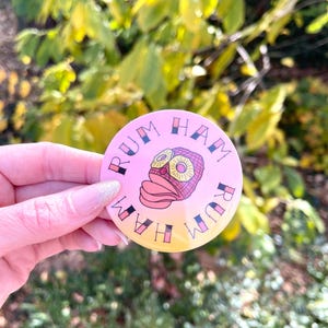 May include: A round, pink button with the words "RUM HAM" encircling a cartoon image of a ham and pineapple. The button has a gradient from pink to yellow. The background is blurred.