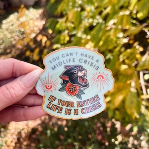 May include: A sticker with a light blue background and a black panther illustration with a red flower. The sticker reads "YOU CAN'T HAVE A MIDLIFE CRISIS IF YOUR ENTIRE LIFE IS A CRISIS" in white and orange text.