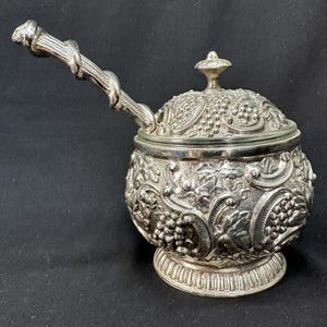May include: Ornate silver-colored serving dish with a lid and handle. The dish features a detailed relief pattern of grapes and leaves. The handle has a twisted vine design. The dish sits on a decorative base, suggesting a vintage or antique style.