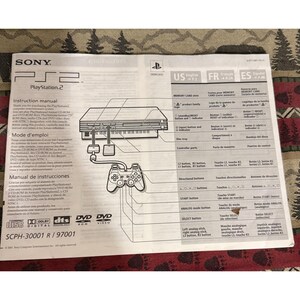 May include: A white Sony PlayStation 2 instruction manual featuring black text and diagrams. The document displays the console and controller layout with labels in English, French, and Spanish, including model number SCPH-30001 R / 97001.