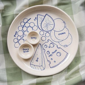May include: A round white ceramic serving plate with two small attached bowls, featuring blue line drawings of grapes, figs, crackers, brie, and swiss cheese labeled with the text grapes, figs, crackers, brie, holy cheese, honey, and Gmas relish.