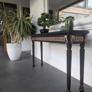 May include: A black console table with gold detailing and four black, fluted legs. The table is decorated with a bonsai tree, a green cactus, and a decorative glass ball. A large potted plant is to the left. The table is approximately 120 cm long.