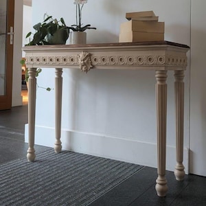 Può includere: Una consolle beige con piano marrone scuro e dettagli intagliati. Il tavolo ha quattro gambe scanalate e un intaglio decorativo a forma di volto sul davanti. Un'orchidea in vaso e libri sono sopra. Il tavolo è lungo 122 cm.