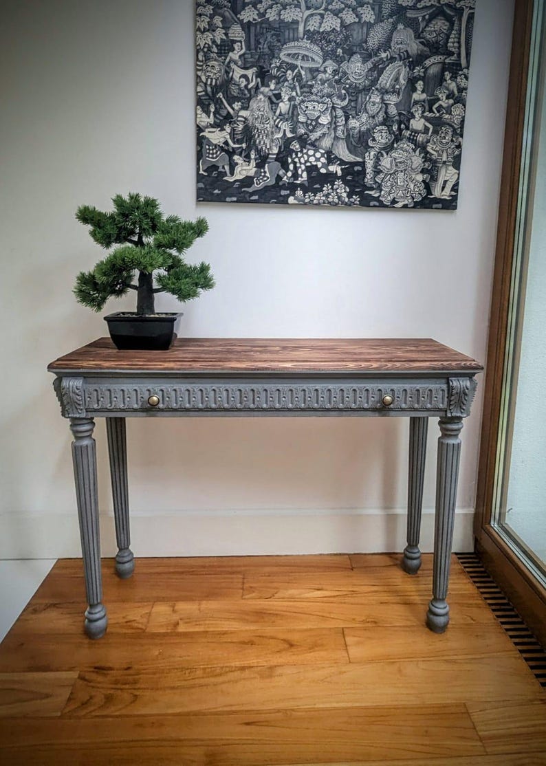 May include: A wooden console table with a grey painted frame and legs. The tabletop is stained brown. A small bonsai tree in a black pot sits on the table. A black and white artwork hangs on the wall above the table.