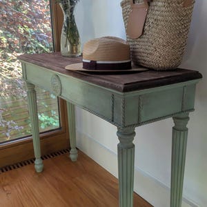 May include: A painted console table with a dark wood top and a pale green base. A straw hat with a burgundy band, a woven basket, and a vase of purple flowers are on the table. The table has fluted legs and rope detailing.