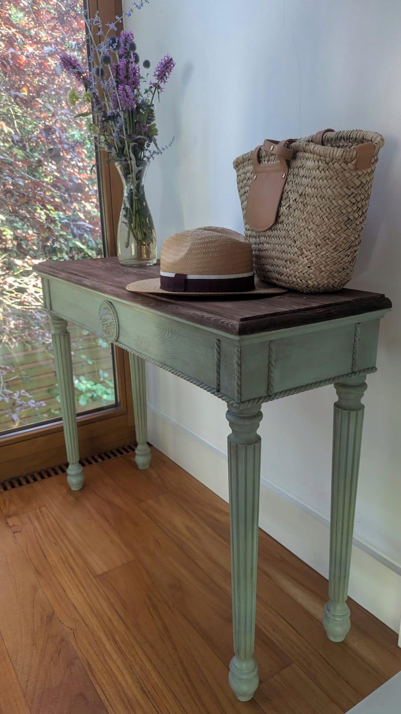 May include: A painted console table with a dark wood top and a pale green base. A straw hat with a burgundy band, a woven basket, and a vase of purple flowers are on the table. The table has fluted legs and rope detailing.