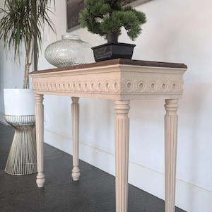 Może przedstawiać: Beżowa konsola z ciemnobrązowym blatem i dekoracyjnymi rzeźbionymi detalami. Stół ma cztery żłobkowane nogi i jest zwieńczony małym bonsai w czarnej doniczce. Na stole stoi szklany wazon.