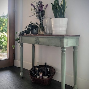 May include: A sage green console table with a dark wood top, featuring a white potted plant, a vase of purple flowers, and a clear glass jug. A wicker basket filled with bottles sits below the table.