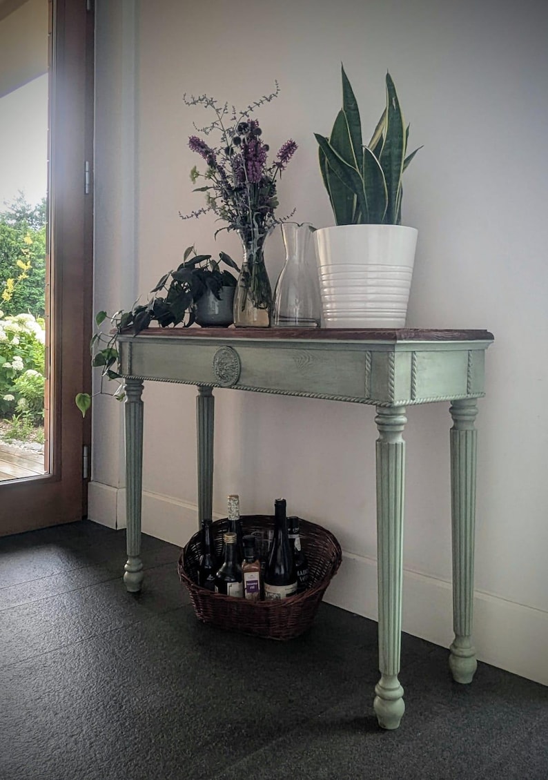 May include: A sage green console table with a dark wood top, featuring a white potted plant, a vase of purple flowers, and a clear glass jug. A wicker basket filled with bottles sits below the table.