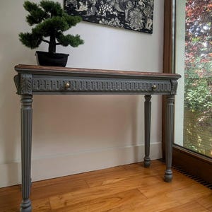 May include: A grey and brown console table with a decorative carved design. The table has four fluted legs and a dark wood top. A small bonsai tree sits on the table. A black and white artwork hangs on the wall above the table.