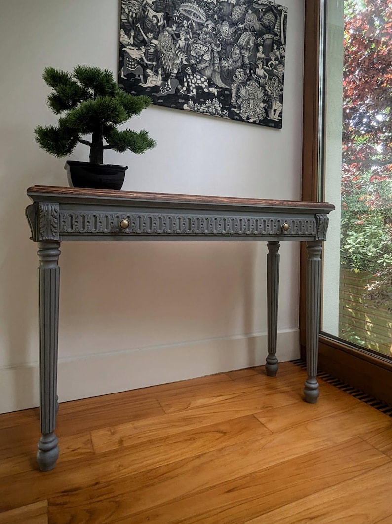 May include: A grey and brown console table with a decorative carved design. The table has four fluted legs and a dark wood top. A small bonsai tree sits on the table. A black and white artwork hangs on the wall above the table.