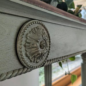 May include: Close-up of a grey painted wooden table with a decorative, circular medallion. The table has a rope-like trim and a dark wood top. The table legs are visible, and the background is blurred.
