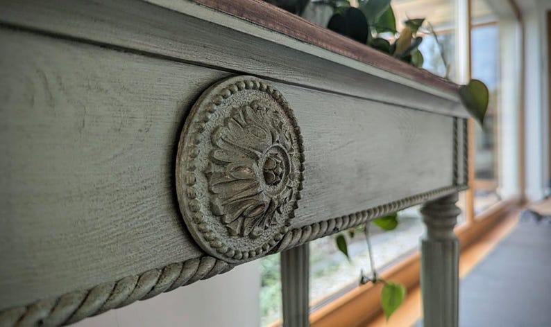 May include: Close-up of a grey painted wooden table with a decorative, circular medallion. The table has a rope-like trim and a dark wood top. The table legs are visible, and the background is blurred.