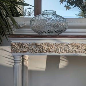 May include: A clear, textured glass vase sits on a decorative white and gold console table. The table features ornate carvings along the front edge and is supported by fluted columns. The background includes a window with greenery and sunlight.