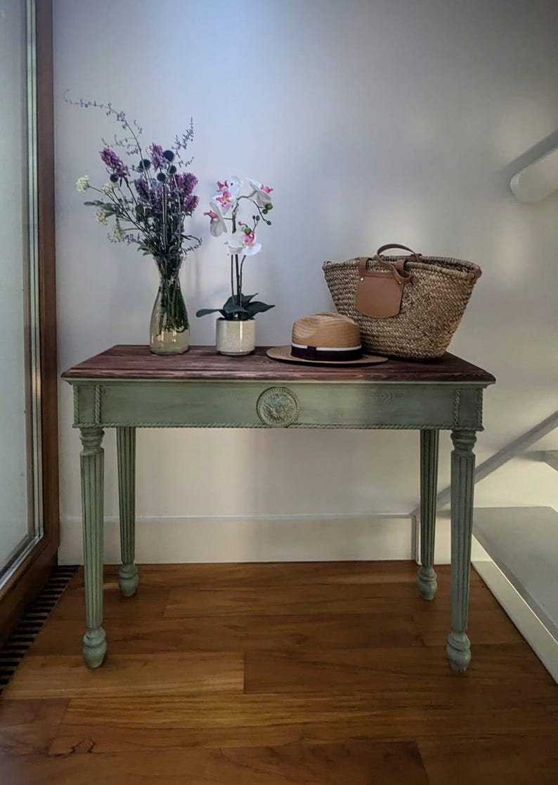 May include: A small, antique-style table with a dark wood top and sage green base. The table is decorated with a straw tote bag, a straw hat, and a vase of flowers. The table's legs are fluted.