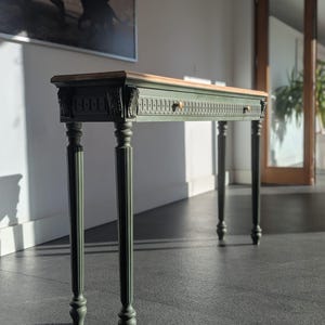 May include: A long, narrow console table with a dark green painted frame and a natural wood top. The table features ornate detailing, two small drawers with brass knobs, and four slender, turned legs. The table is approximately 122 cm long.