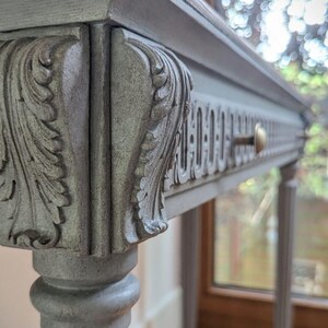 May include: A grey painted console table with ornate carved details. The table features a decorative drawer with a brass knob and is supported by slender, fluted legs. The top is a darker wood. The table is a classic piece of furniture.
