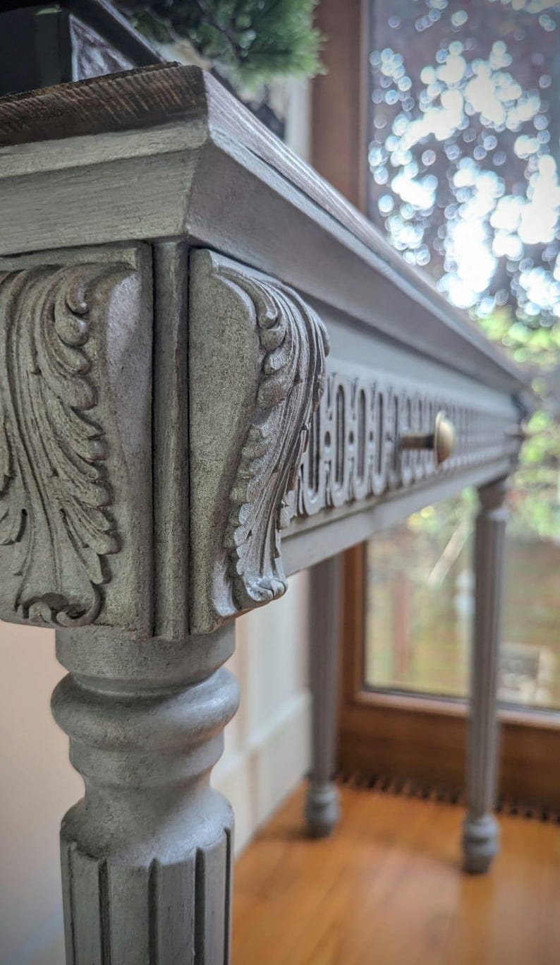 May include: A grey painted console table with ornate carved details. The table features a decorative drawer with a brass knob and is supported by slender, fluted legs. The top is a darker wood. The table is a classic piece of furniture.
