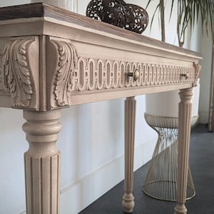 Può includere: Una consolle beige chiaro con dettagli intagliati e un piano marrone scuro. Il tavolo presenta due cassetti con pomelli in ottone ed è sostenuto da gambe scanalate. Sulla parte superiore del tavolo sono presenti dei globi decorativi.